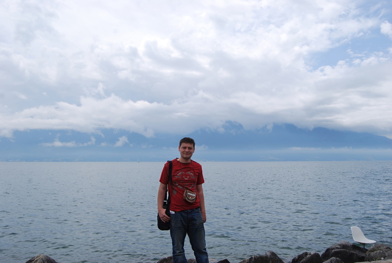 9 - A man wearing a red t-shirt and blue jeans standing on a rocky shore in front of a large body of water with distant mountains under a cloudy sky